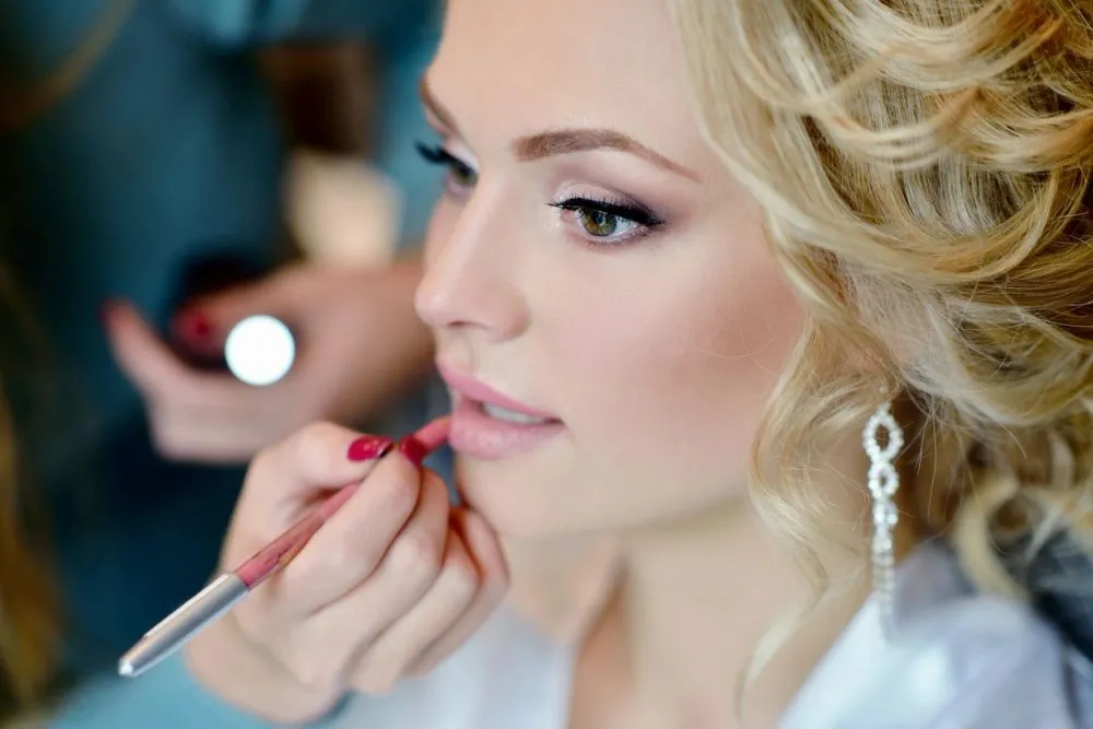 A stylist applying lip color for a bride with curled blonde hair
